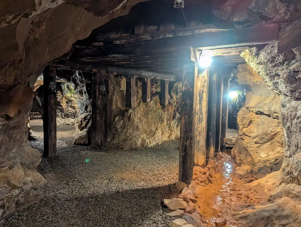 Underground gold-mine tunnel with rough rock walls, timber posts and an iron-stained water channel